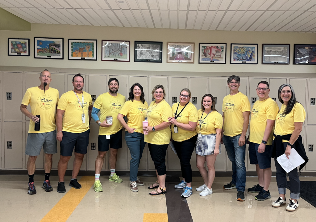 A group of ten people in yellow shirts stand in front of lockers, smiling and posing in a school hallway.